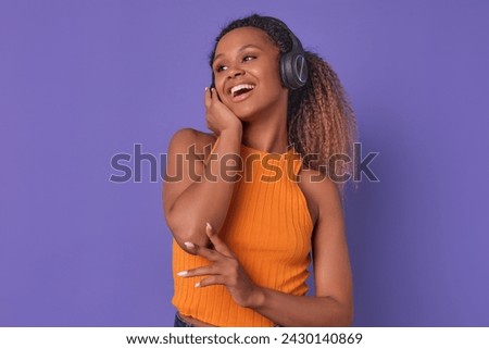 Young laughing ethnic African American woman in headphones listens to humorous podcast from famous comedians running audio blog on internet posing on isolated purple background. Music, songs