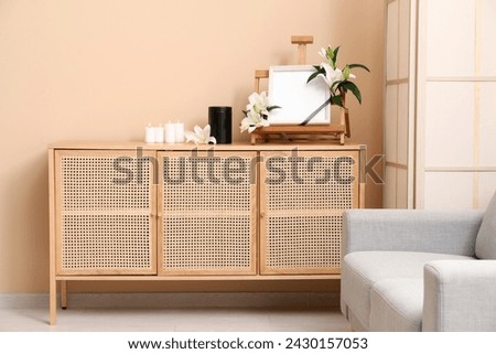 Blank funeral frame with lily flowers, mortuary urn and burning candles on wooden cabinet against color wall Royalty-Free Stock Photo #2430157053