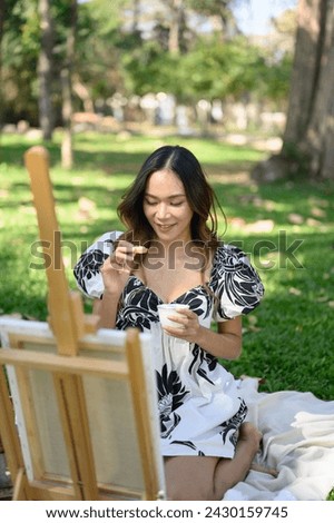 Happy young woman painting on easel sitting on the grass during a picnic in the park