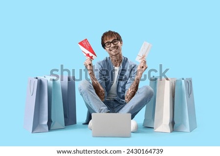 Young tattooed man with gift cards, laptop and shopping bags on blue background