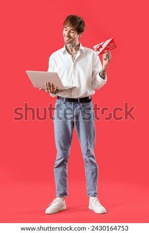 Young tattooed man with gift voucher and laptop on red background