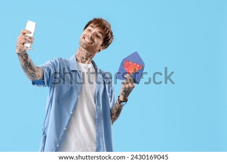 Young tattooed man with gift card taking selfie on blue background