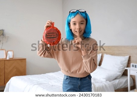 Funny little girl in wig with whoopee cushion showing silence gesture at home. April Fools' Day celebration Royalty-Free Stock Photo #2430170183