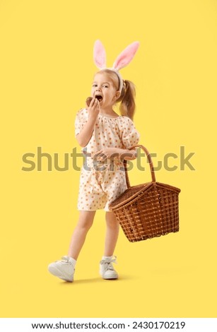 Cute little girl in bunny ears with basket eating chocolate Easter egg on yellow background Royalty-Free Stock Photo #2430170219