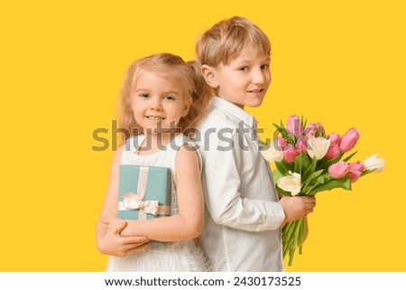 Cute little children with bouquet of beautiful tulips and gift box on yellow background. International Women's Day