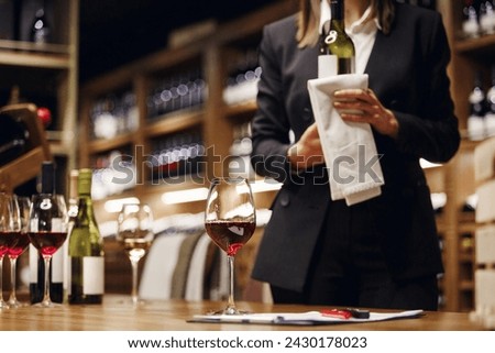 Poured glass of red wine against the background of the interior of a restaurant or cellar with a sommelier standing behind with a bottle of elite alcohol drink. Royalty-Free Stock Photo #2430178023