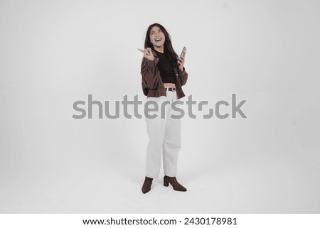 Smiling young Asian girl wearing brown jacket is standing while holding her phone and pointing to a copy space beside her. Royalty-Free Stock Photo #2430178981