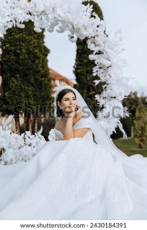 A brunette bride in a veil and a tiara in her hair sits on a spread dress, posing near a white arch made of flowers in the shape of a circle. On a white background. Sunny day. Wedding ceremony