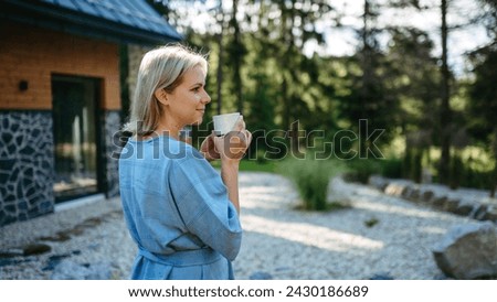 Happy young woman standing in garden near cottege and enjoying cup of morning coffee on summer vacation in mountains.