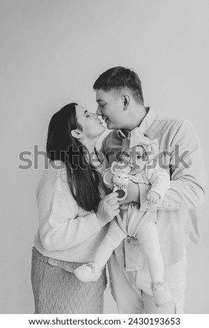 Father kiss mother hugging daughter. Happy family hugs cute baby on holiday. Dad, mom holds hands infant. Daddy, mommy embrace girl with bow in hair isolated on wall. Black and white photo. Family.