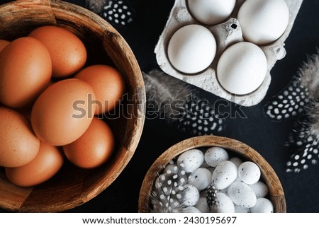 Brown eggs in a wooden bowl, white chicken eggs in a tray next to quail eggs and feathers on a dark background