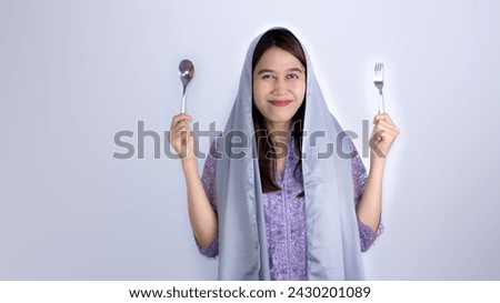 Excited and hungry young Asian Muslim woman holding spoon and fork, expecting delicious healthy food after fasting in ramadan