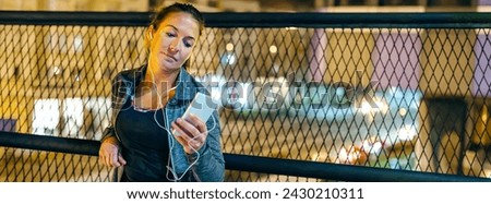 Banner of young woman runner listening music on mobile phone application on evening. Portrait of female athlete with earphones resting over bridge banister after training on town. Right copy space.