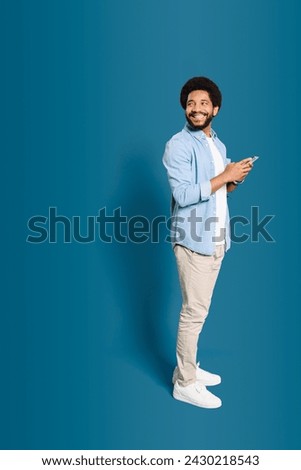 A cheerful Brazilian man in casual wear using smartphone standing in full length isolated on blue, looking back at copy space suggesting a place for advertising, male freelancer or student with phone