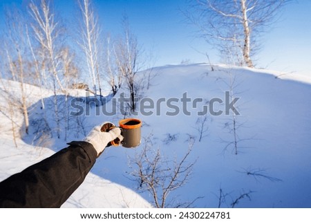 Hiking mug in the hand of a tourist against the background of a winter forest, drinking tea in the mountains in the cold, white snow, snowdrifts, a glass for hot drinks. High quality photo
