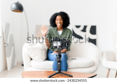 A vivacious African-American vlogger warmly greets her online community, ready to start a dynamic video blog session from her minimalist, yet trendy living space Royalty-Free Stock Photo #2430234555