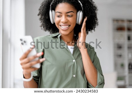 A vivacious African-American woman wearing headphones enjoys a lively video call, her smile illuminated by the daylight streaming through a window in a chic, minimalist office setting. Royalty-Free Stock Photo #2430234583