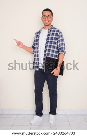 Full body portrait of man carry a laptop pointing to the right side with happy expression  Royalty-Free Stock Photo #2430264903