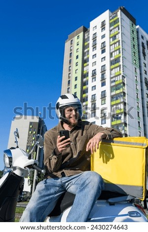A courier with a phone is waiting for his order. A man with a yellow box and on a scooter outside