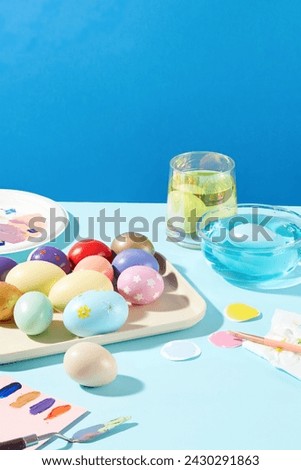 Greetings and presents for Easter with a tray featured lots of Easter eggs in variety colors and patterns. A brush, a palette knife and paint colors are displayed with a cup and bowl of color water