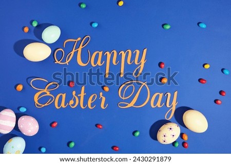 Some letters made of yellow paper combined form the text Happy Easter Day against the blue background. Lots of candies and Easter eggs decorated. Easter poster and banner template