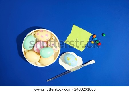 Happy Easter Day concept with a white dish containing many Easter egg painted in pastel colors arranged with colorful candies and a brush. A note paper with copy space for Easter content