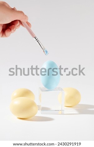An Easter egg in blue color is placed on a glass podium in cube shaped, surrounded by other yellow eggs. A hand holding a brush to paint the egg on the podium. Easter means the celebration of hope