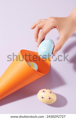 A rolled paper in orange color decorated with few Easter eggs painted with flowers and stripes. A female hand is holding a pastel blue egg. Easter Day is a special day for Christians