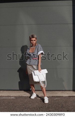 Street style, fashion: elegant woman wearing trendy stripped oversize t-shirt, sequin bright midi skirt, holding leather bag, posing in street of city over grey wall. Copy, empty space for text
