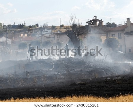 A firefighter puts out the fire with a hose along with another firefighter amidst the smoke of a fire on the dry ground of a field.