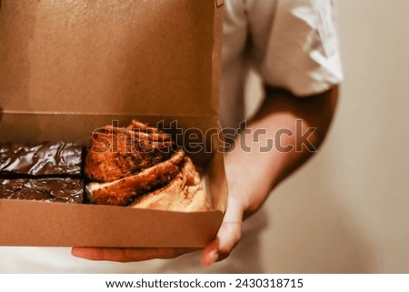 Hands holding a craft box with buns close up. Delicious cinnamon pastries and chocolate brownies. Fresh baked goods delivery concept Royalty-Free Stock Photo #2430318715