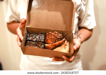Hands holding a craft box with buns. Delicious cinnamon pastries and chocolate brownies. Fresh baked goods delivery concept Royalty-Free Stock Photo #2430318757