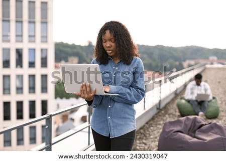 Smiling female using laptop outdoors, male colleague works sitting in bag chair in background. Modern lifestyle, connection, business, communication, web chat, social media, video conference concept.