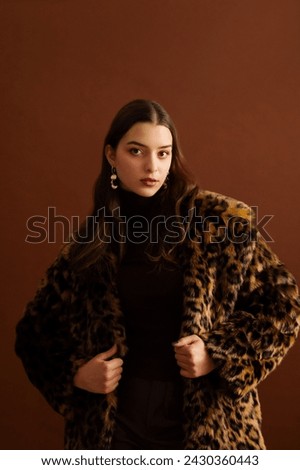 Fashionable confident woman wearing hat, trendy faux fur leopard print coat, posing on brown background. Studio fashion portrait. Copy, empty, blank space for text
