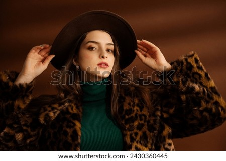 Fashionable confident woman wearing hat, trendy faux fur leopard print coat, green turtleneck, posing on brown background. Studio fashion portrait. Copy, empty, blank space for text
