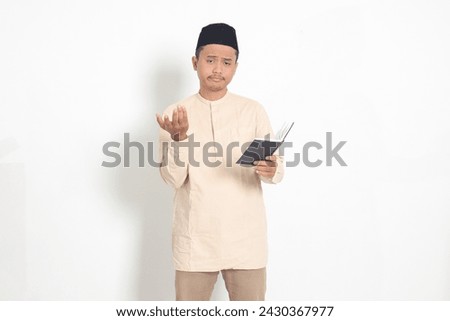 Portrait of confused Asian muslim man in koko shirt with peci difficulty understanding the contents of the book, reading a textbook. Isolated image on white background Royalty-Free Stock Photo #2430367977