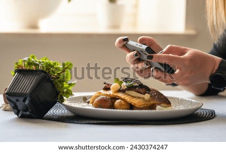 A woman taking photo with smart phone of the food. Roasted branzino with marinated mushrooms and butter potatoes on the plate. Posting and sharing food pictures on social media. Lifestyle.