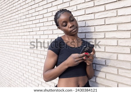 This image features a young African woman engrossed in her smartphone while standing against a white brick wall. Her posture is relaxed, and she holds the phone with both hands, indicating she is