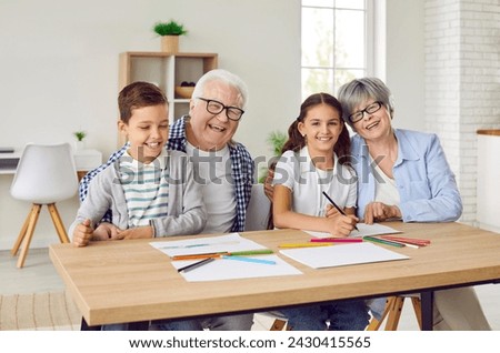 Happy grandparents and children drawing together. Group family portrait of cheerful grandmother, grandfather and little children sitting at table with paper and pencils, looking at camera and smiling