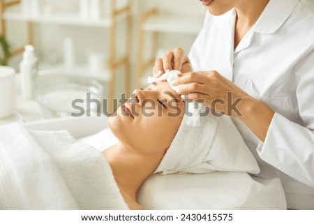 Close-up photo pf professional cosmetologist or beautician doctor hands making anti-age procedures cleaning with napkins facial mask of young pretty woman client in beauty spa clinic. Royalty-Free Stock Photo #2430415575