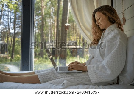 A woman enjoys the tranquility of working remotely from a cozy nook, wrapped in a waffle robe with a laptop, surrounded by forest views. The serene environment complements her focused demeanor. Royalty-Free Stock Photo #2430415957