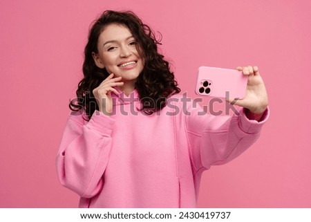happy smiling curly woman in pink hoodie posing on pink studio background holding smartphone using device spring summer fashion style taking selfie photo on camera