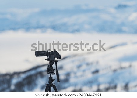 Video camera on tripod stands high in mountains filming remote winter hills. Closeup focus on camera with blurry background