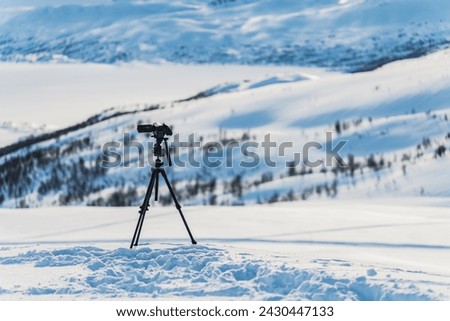 Video camera on tripod stands high in mountains filming remote winter hills. Focus on camera with blurry background