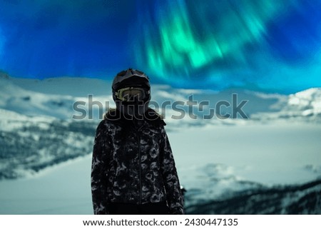 Snowmobile driver on sport helmet stands high with winter mountains, frozen lake and Aurora Borealis green lights in sky as a blurry background. close up photo. Women in winter jacket