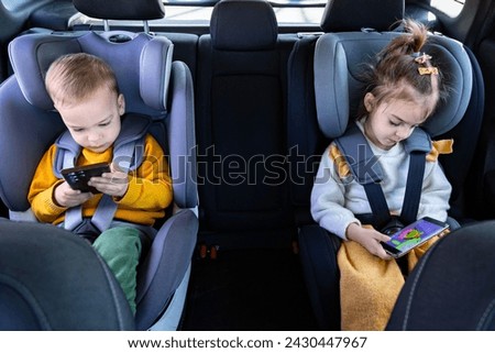 Front view of brother and sister on car back seat holding smart phones while traveling.