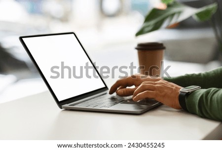 Cropped of african american guy sitting at table next to window, drinking coffee, typing on laptop computer with white blank screen, working online, cafe interior, mockup, copy space