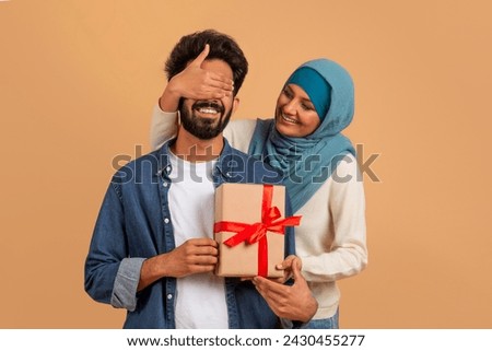 Surprise for you. Romantic muslim woman covering husbands eyes and giving present, greeting him with birthday, wedding anniversary or valentines day, standing on beige studio background