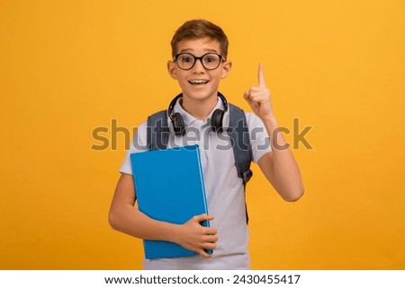 Excited Teen Boy Wearing Eyeglasses Having Idea, Raising Finger Up, Clever Teenage Male Child Holding Books, Ready To Answer A Question, Standing On Yellow Studio Background, Copy Space