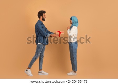 Smiling arab man in denim shirt presenting surprise gift box to excited muslim wife while they standing together against beige studio background, romantic husband greeting spouse with valentines day
